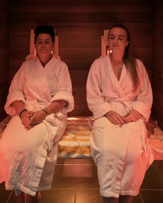 Due donne si rilassano in sauna a infrarossi con accappatoi bianchi e occhi chiusi